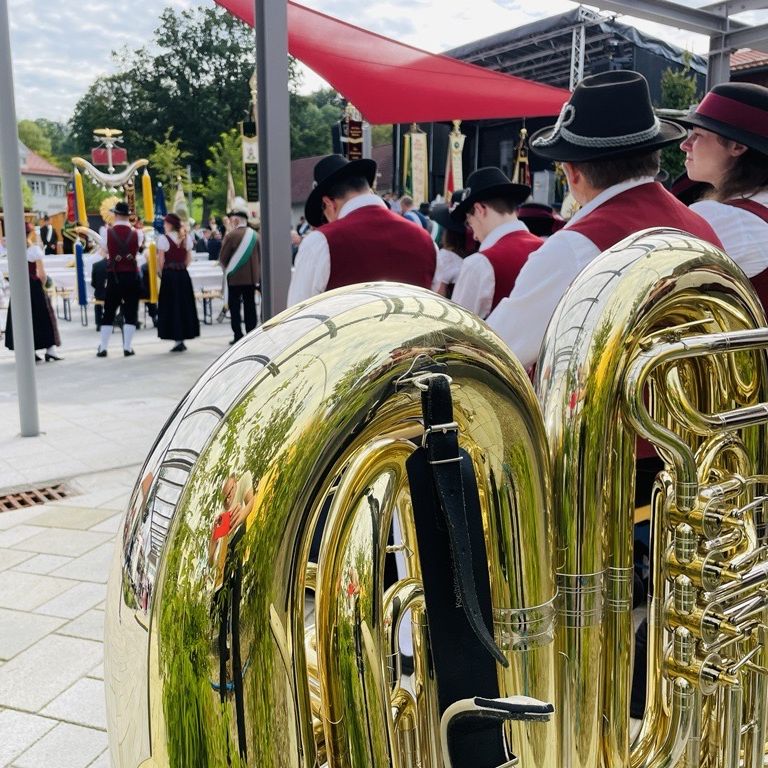 Im Vordergrund des Bildes stehen zwei Tuben auf dem Boden. Im Hintergrund sind Musikanten und Besucher der Dorfplatzeinweihung zu erkennen.